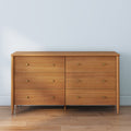 Mid-century modern six-drawer natural wood dresser with brass knobs on hardwood floor against pale blue wall, bedroom storage furniture