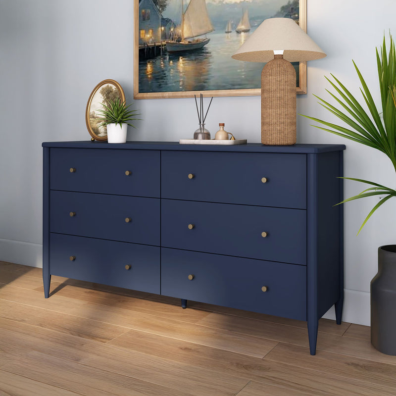 Navy blue six-drawer dresser with brass knobs in modern bedroom, styled with a woven lamp, potted plants and a nautical framed painting.