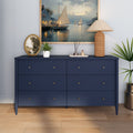 Navy blue six-drawer dresser with brass knobs on hardwood floor, styled with framed sailboat painting, wicker table lamp, decorative tray and potted plants — modern coastal home decor