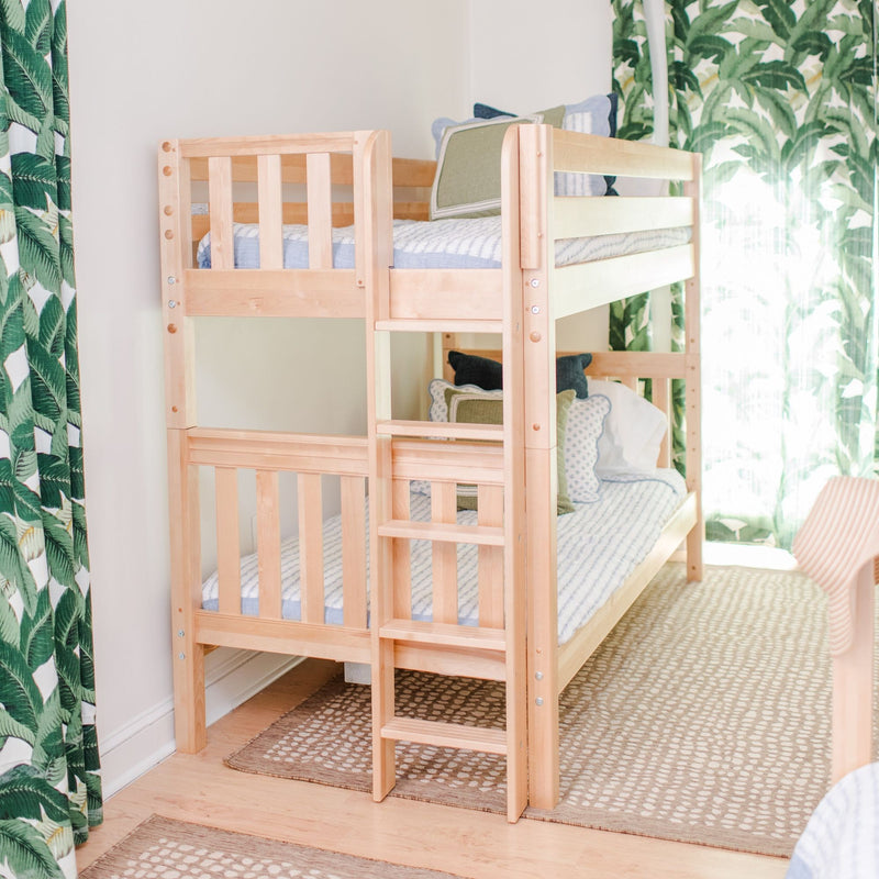 Light wood bunk bed with ladder in a bright kids' bedroom, blue-green bedding, tropical leaf curtains and patterned rug, modern children's room décor