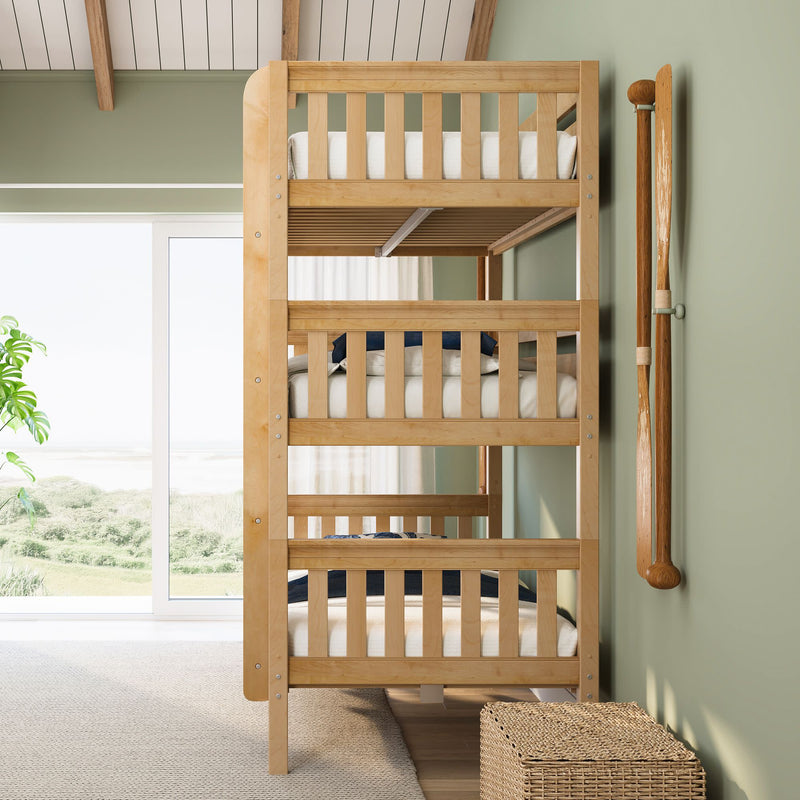 Light wood triple bunk bed in sunlit coastal bedroom with sage green walls, sliding glass door showing dunes and ocean view, wicker storage basket and wooden oar decor