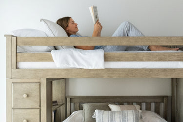 Woman reading on top bunk of natural wood loft bunk bed with drawers and cozy pillows in a bright neutral bedroom.