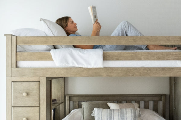 Woman reading on top bunk of natural wood loft bunk bed with drawers and cozy pillows in a bright neutral bedroom.