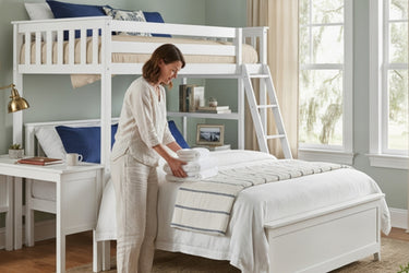 Woman making bed in bright coastal-style bedroom with white twin-over-full bunk bed and ladder, white linen bedding and blue accent pillows