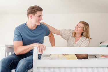 Smiling young couple sitting on a white bed in a bright cozy bedroom, happy homeowners enjoying their new home.