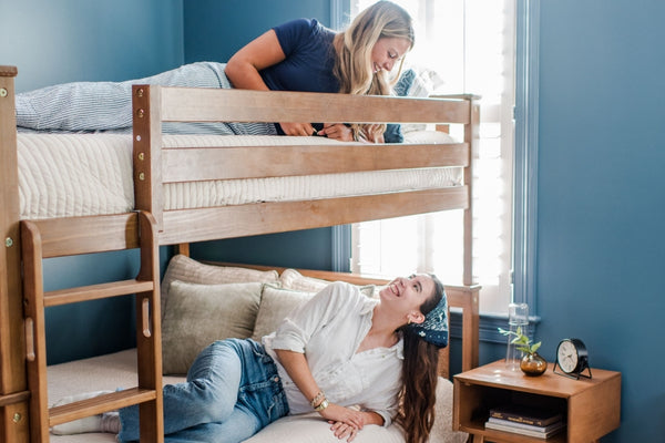 Two people smiling on a wooden bunk bed in a cozy blue bedroom with natural light, bedside table and plant.