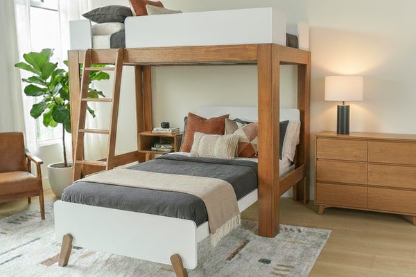 Space-saving wooden loft bed with lower twin, ladder, neutral bedding, dresser, lamp and potted plant in a modern bedroom.