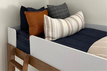 White kids loft bed with wooden ladder, navy quilted bedding and decorative pillows — striped lumbar pillow, brown leather throw pillow and gray plaid cushion in a modern minimalist bedroom.