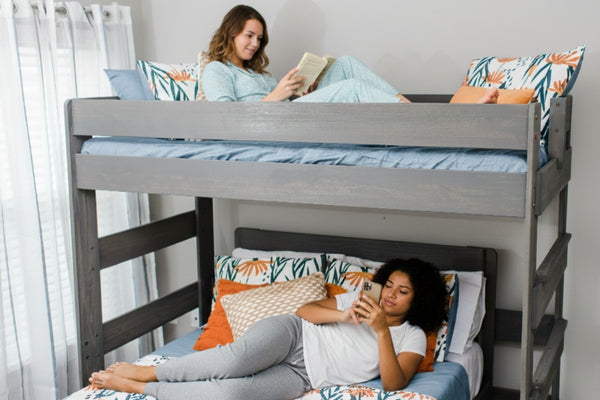 Two women relaxing on a gray wooden bunk bed in a bright modern bedroom, top bunk reading a book and bottom bunk using a smartphone with colorful patterned bedding.