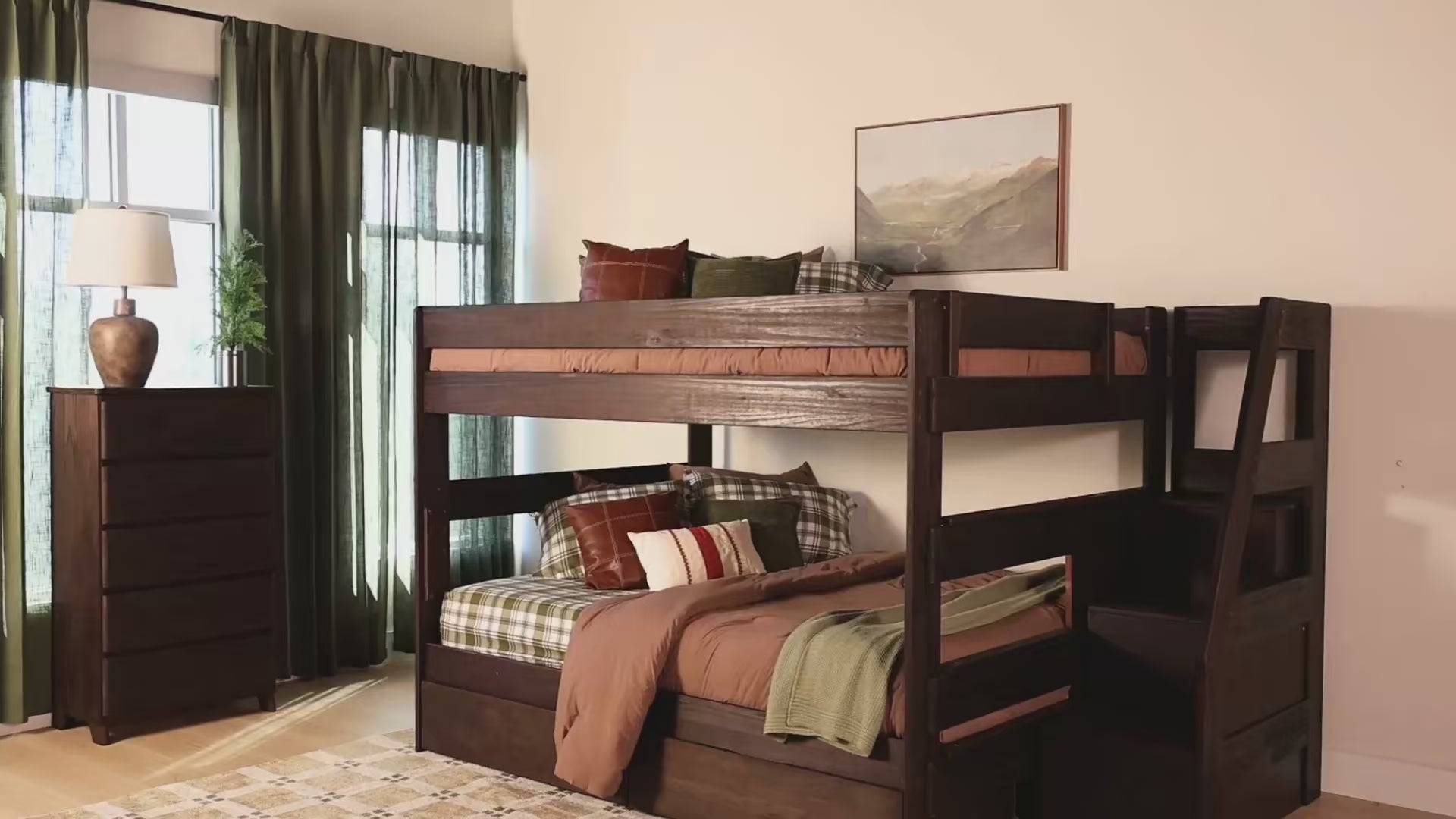 Two women in cozy sweaters and jeans relax and play on a dark wood twin-over-twin bunk bed with a stair unit, plaid bedding, and a built-in storage drawer in a warm, softly lit bedroom.