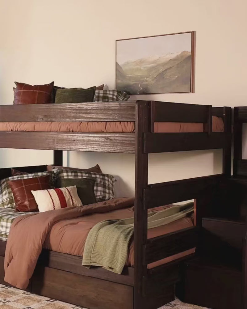 Two women in cozy sweaters and jeans relax and play on a dark wood twin-over-twin bunk bed with a stair unit, plaid bedding, and a built-in storage drawer in a warm, softly lit bedroom.